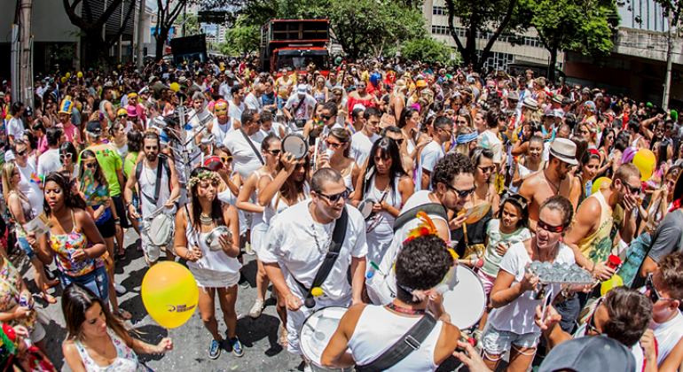 Membros de um bloco de carnaval toca instrumentosmusicais; um pouco atrás, mais de trezentas pessoas acompanham; mais ao fundo há a presença de um caminhão de som.