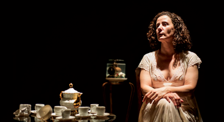 Cena de peça teatral com mulher sentada de vestido com semblante contemplativo; a seu lado, um jogo de chá.