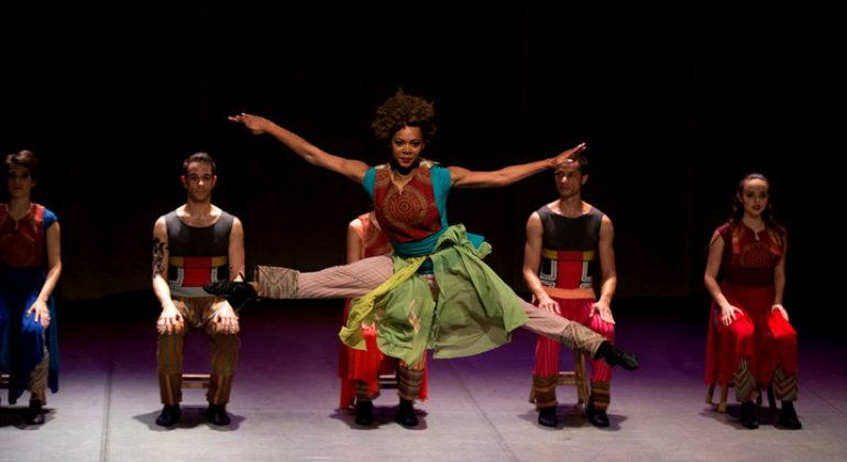 Mulher dá um pulo de dança em apresentação cênica, atrás, quatro pessoas estão sentadas.
