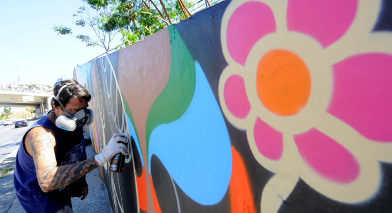 O prazo de inscrições de propostas de arte urbana para o Concurso Gentileza foi prorrogado até o dia 7 de fevereiro. Foto: Rodrigo Clemente/PBH Homem com máscara de segurança e luva grafita muro com flores e cores vibrantes, durante o dia.