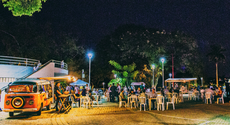 Praça da Pampulha mostra food truck e pessoas sentadas em mesas, conversando. 