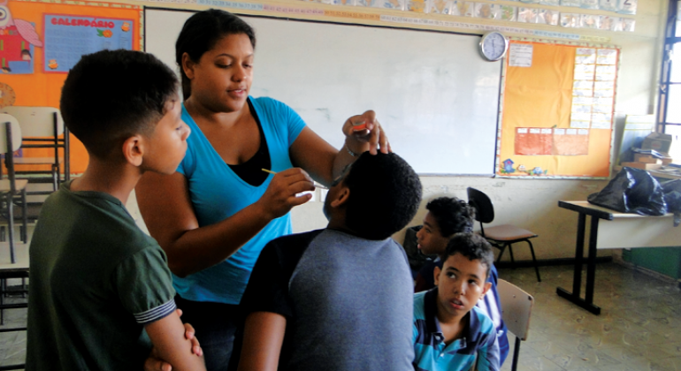 Foto: Divulgação PBH Monitora do Programa Escola nas férias faz pintura facial em criança, outros três garotos observam e aguardam.