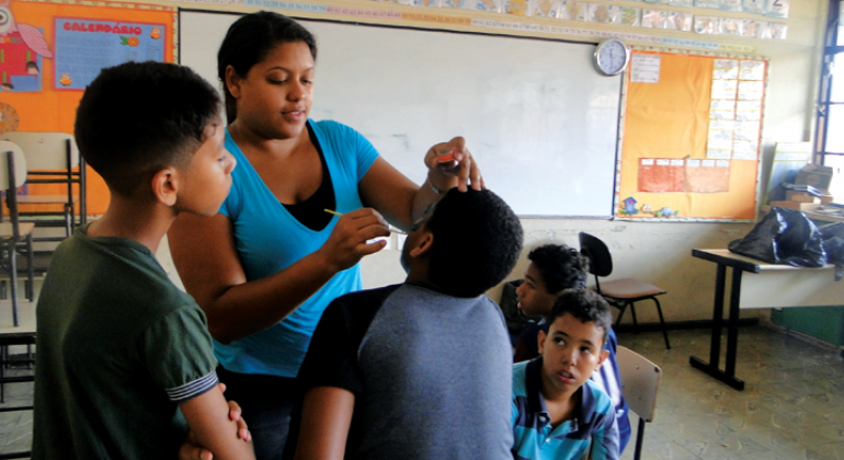 Monitora do Programa Escola nas férias faz pintura facial em criança, outros três garotos observam e aguardam. 