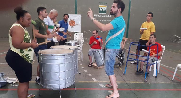 Homem comanda grupo de quatro percussionistas; ao lado, duas pessoas com dificuldade de locomoção assistem, sentadas; uma está de pé com chocalho.