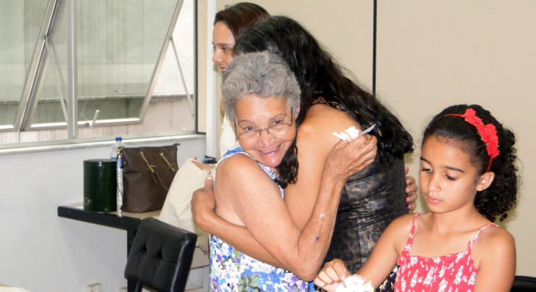 Duas mulheres se abraçam, uma menina está mais à frente e à direita e uma mulher, atrás e à esquerda.