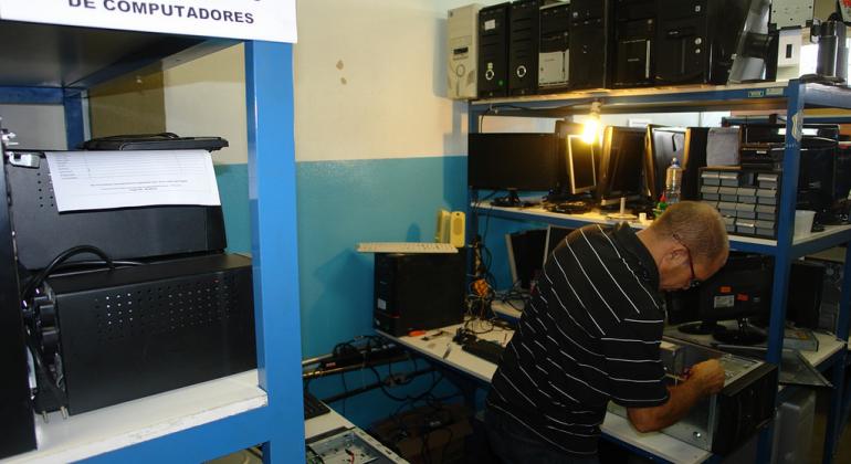 Técnico recupera computadores em sala com diversos componentes. Em uma estante à frente à esquerda, há uma placa com os dizeres: Recondicionamento de computadores. 