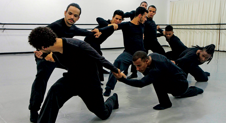 Oito homens vestidos de preto interagem em apresentação de dança.