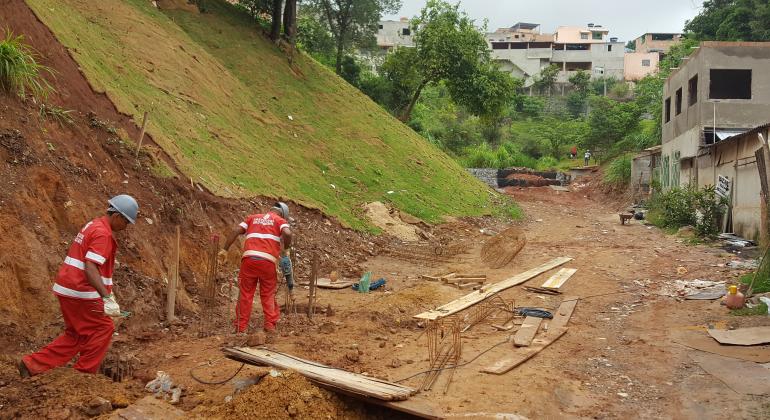 PBH faz obras de eliminação de risco geológico no Barreiro Dois trabalhadores com uniformes da Prefeitura executam obra em encosta no Barreiro. Ao lado, há construções inacabadas e a rua está sem capeamento.