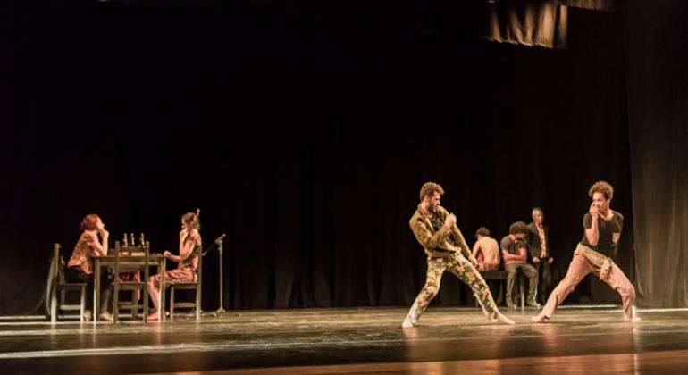 Atores dançando no palco; ao fundo, duas pessoas estão sentadas em uma mesa.