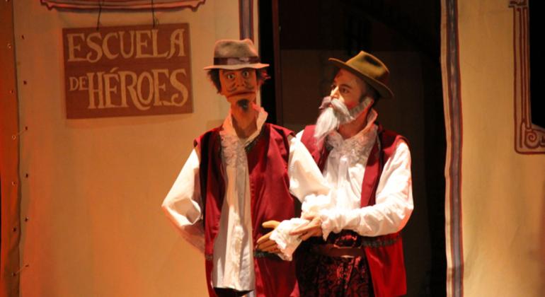 Personagem de peça teatral, Dom Quixote tem um boneco na mão direita; na parede do cenário ao fundo vê-se uma placa em que está escrito os dizeres "Escola de Heróis".