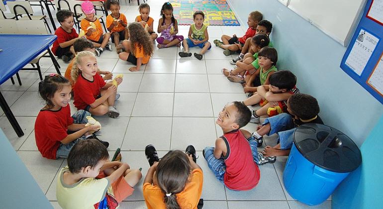 Cerca de 18 crianças sentadas em círculo em sala de aula.