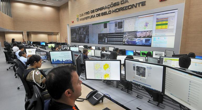 Mais de vinte pessoas em três fileiras, cada uma delas em frente ao computador; à frente, um painel grande de vídeo em uma parede. No alto os dizeres: Centro de Operações da Prefeitura de Belo Horizonte. 