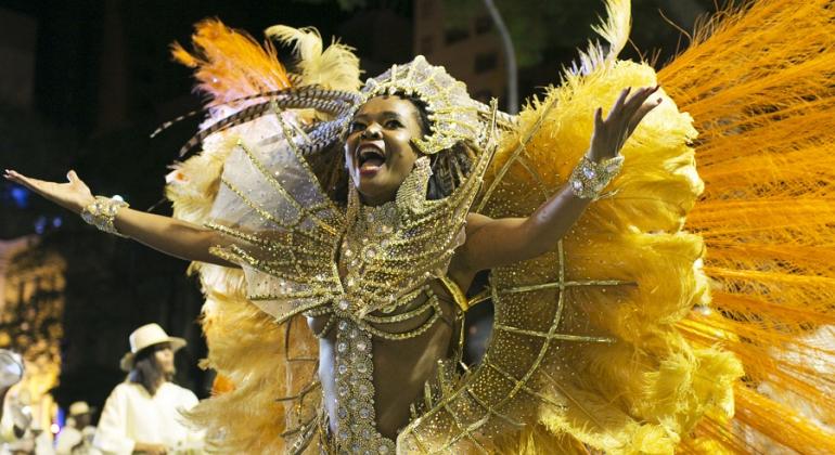 Passista de escola de samba, com adereços dourados, amarelos e laranjas, desfila no chão, à noite. 