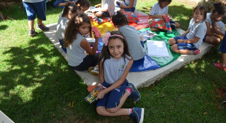 Criança sorri sentada, ao lado de seis colegas, lendo, sentados no jardim, durante o dia.