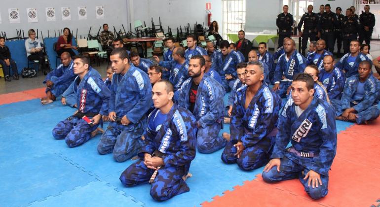 Vinte e um guardas municipais de uniforme azul, ajoelhados, em cerimônia de troca de faixa de Jiu-Jitsu