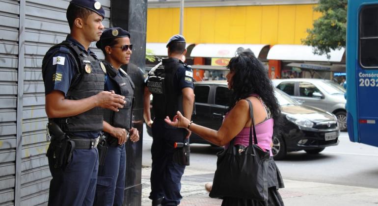 Dois guardas municipais conversam com cidadã na rua, um terceiro guarda observa a rua.