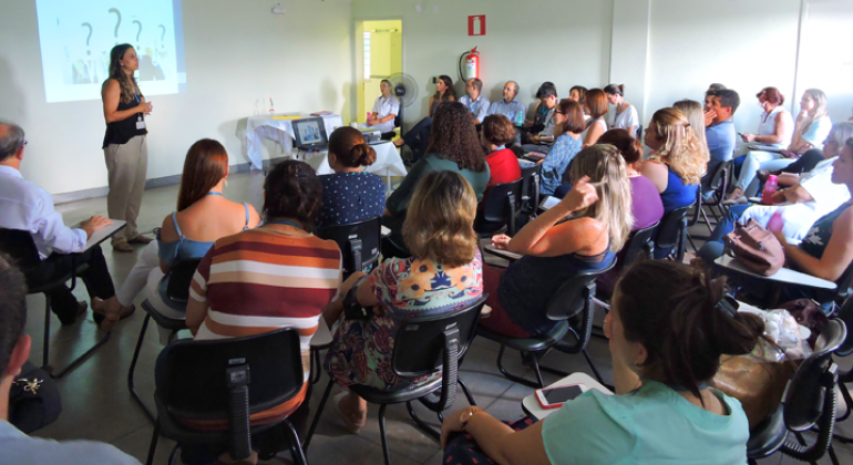 Instrutora apresenta trabalho de Grupo de Trabalho de Promoção da Saúde na Pampulha em uma sala com mais de 50 pessoas. 