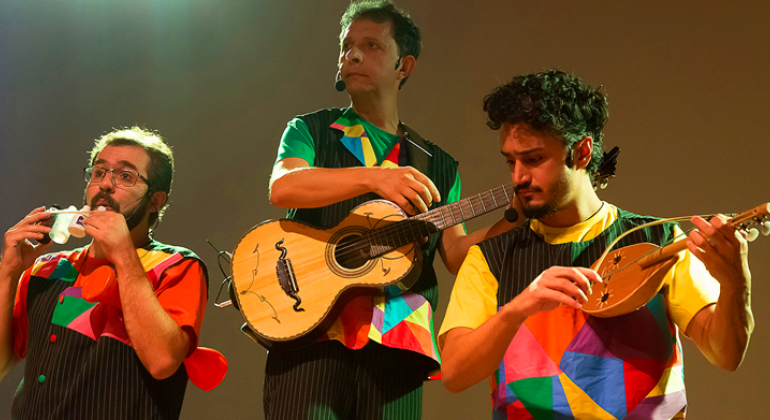 Foto: Guto Muniz Três homens se apresentam com camisetas coloridas e instrumentos musicais.