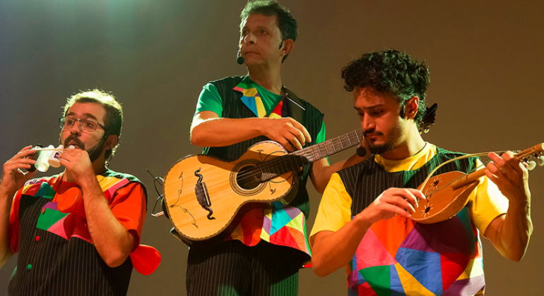 Três homens se apresentam com camisetas coloridas e instrumentos musicais.