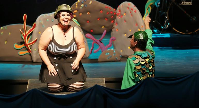 Teatro Francisco Nunes exibe peça infantil com canções que chamam a atenção para a importância da água e dos seres que nela vivem. Foto: Divulgação Atriz com roupa estilizada de balho conversa com homem fantasiado de jacaré, vestido de verde com escamas em cenário teatral aquático.
