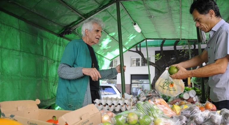 Entre frutas, biscoitos a roscas, programa da PBH leva para a população pontos de comercialização da agricultura familiar. Foto: Rodrigo Clemente/PBH Homem vende legumes e ovos a cidadão em banca de feira.