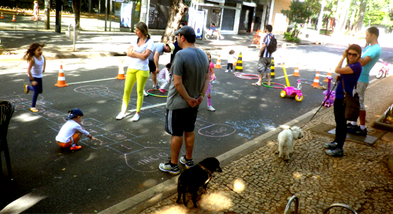 crianças brincam de desenhar no chão e de amarelinha em rua fechada da Savassi, acompanhadas por adultos, durante o dia. 