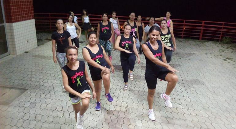 Quinze mulheres de diferentes idades fazem passo de zumba, com uma das pernas levantadas