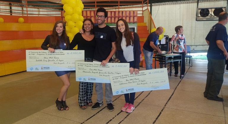 Quatro vencedores de concurso de vídeo "O Trânsito e o Valor da Vida", promovido pela BHTrans, exibem três cheques grandes.