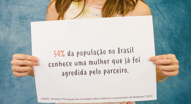 Mulher segura cartaz com a frase: "54% da população no Brasil conhece uma mulher que já foi agredida pelo parceiro" em ação no HMDCC