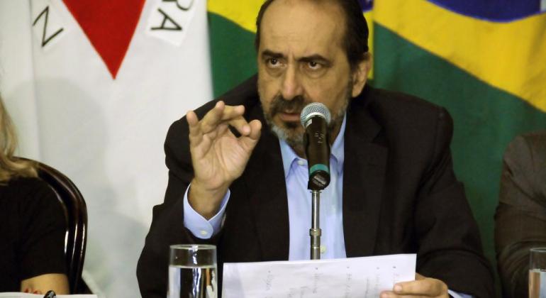 Kalil apresenta balanço do primeiro ano de gestão na Câmara Prefeito Alexandre Kalil segura papel e fala ao microfone. Ao fundo, as bandeiras do Brasil e de Minas Gerais