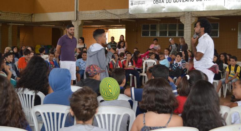 Três pessoas de pé, sendo dois adultos e um jovem, no meio de um círculo formado por dezenas de jovens sentados, em escola