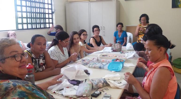 Cerca de nove mulheres fazem bordados em torno de uma mesa grande.