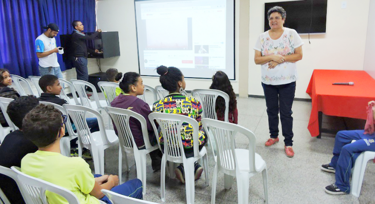 sete crianças assistem a exposição de professora em sala com projetor multimídia; à esquerda, dois monitores operam projetor.