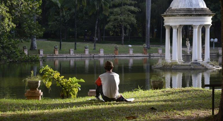 Cidadão, sentado na grama, observa lago e coreto do Parque Municipal Américo Renné Gianneti.