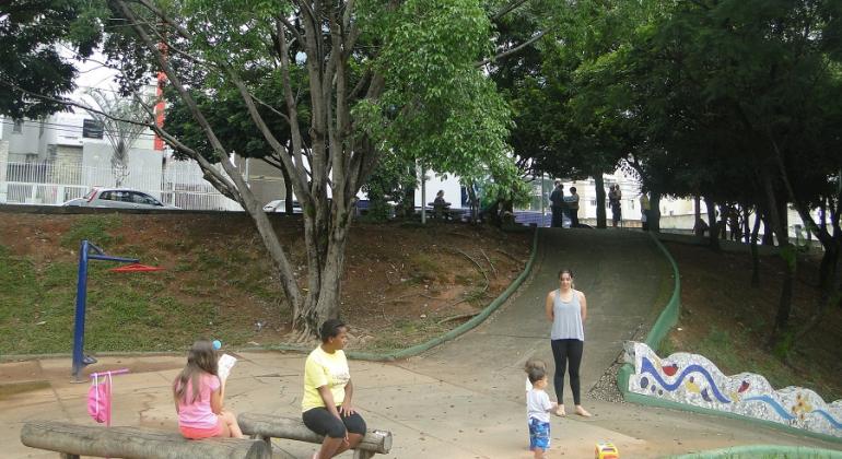Praça Vereador Marco Antônio de Menezes é local de lazer, prática de atividades físicas e de esportes, brincadeiras e contemplação. Foto: Divulgação PBH Três crianças e dois aduntos na praça Vereador Marco Antônio de Menezes, durante o dia.