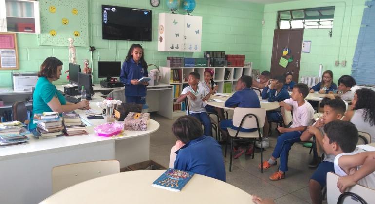 Aluna lê livro em sala de aula cheia, com professora e mais de dez alunos.