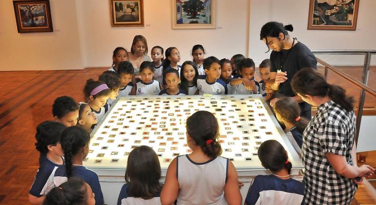 Mais e trinta crianças observam peça de museu, acompanhados por mediador.