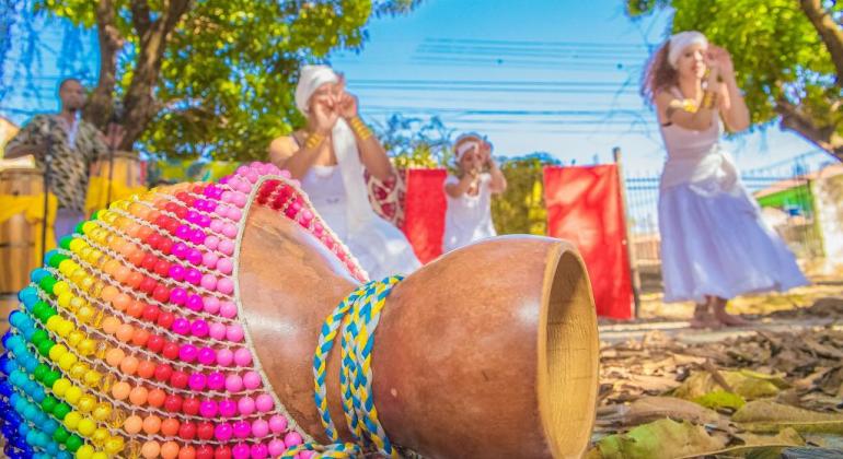 Cia Motumbá se apresenta. Em primeiro plano, há um instrumento musical feito de cabaça. Ao fundo, grupo de mulheres vestidas de branco dançam.