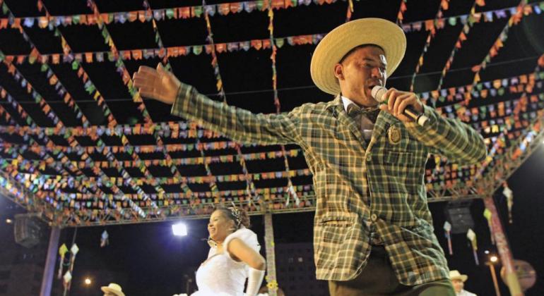 Edital selecionará artistas de forró e sertanejo para compor programação musical dos festejos juninos. Foto: Amira Hissa/PBH Homem com chapéu de palha e blazer xadrez, tem microfone na mão; ao fundo, mulher vestida de noiva e dois outros integrandes de quadrilha junina, no céu, bandeirolas.