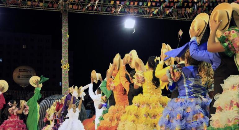 Grupo de dançarinos de quadrilha se apresenta na Praça da Estação de Belo Horizonte. Os homens usam camisas xadrez e as mulheres usam vestidos coloridos e rodados.