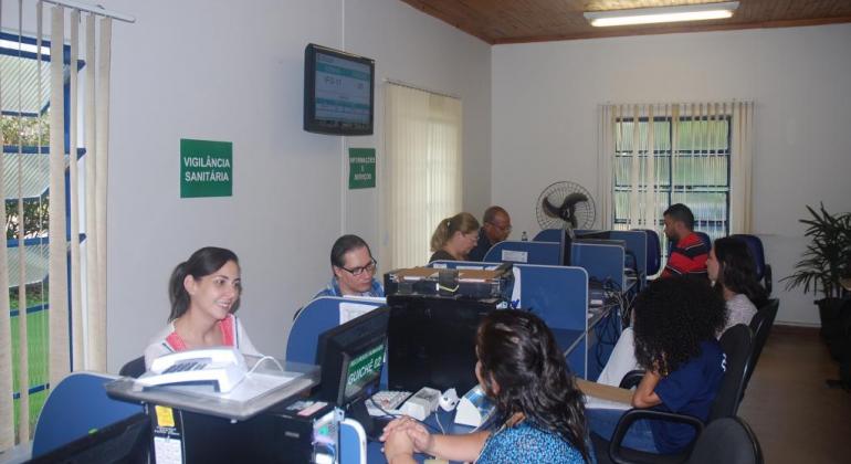 Sala com quatro atendentes sentados em guichês com computadores atendendo cidadãos.