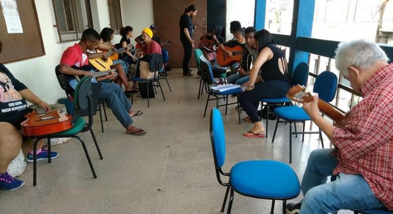 Professor, de pé dá aula de violâo para nove alunos, sentados. 