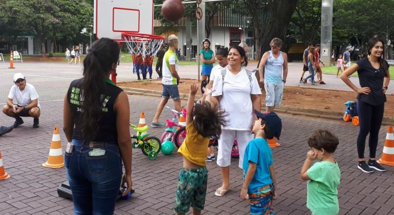 Quatro crianças jogam basquete, acompanhadas por dois adultos, em avenida fechada.