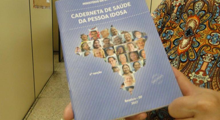 Idosa segura Caderneta de Saúde da Pessoa Idosa. Na capa da caderneta, há um mapa do Brasil com montagem de fotos de pessoas idosas.