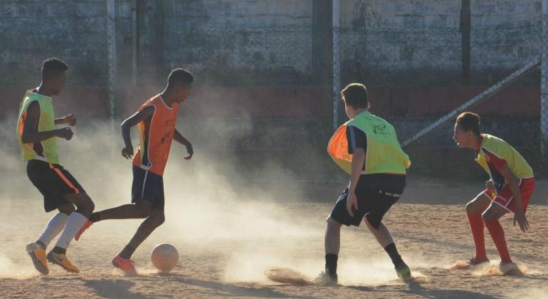 Quatro atletas do futebol na foto. Três deles usam colete verde e um, com a bola no pé, usa colete laranja.