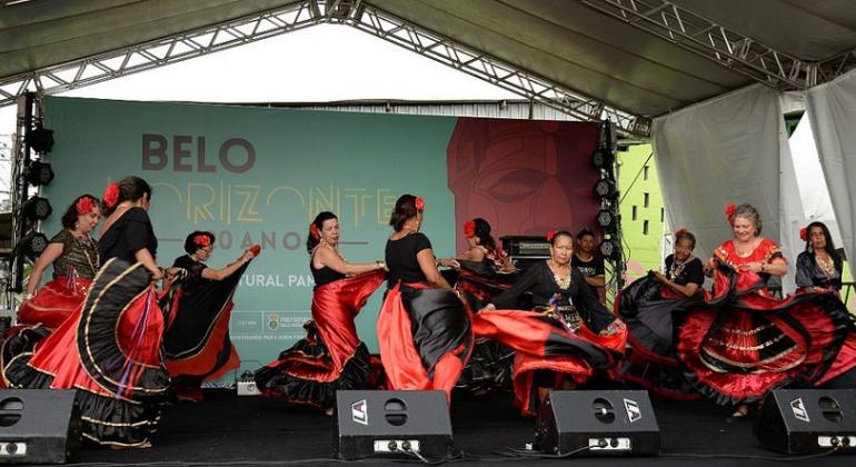 Nove mulheres vestidas de preto e vermelho fazem apresentação de dança cigana em palco. 