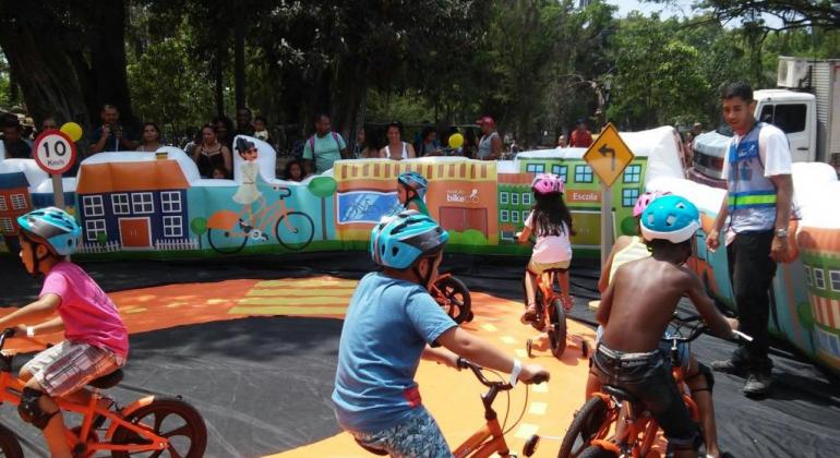 Quatro crianças em biciletas, auxiliadas por monitor, em Escolinha de Bike circuito didático de trânsito, na Fundação de Parques Municipais.
