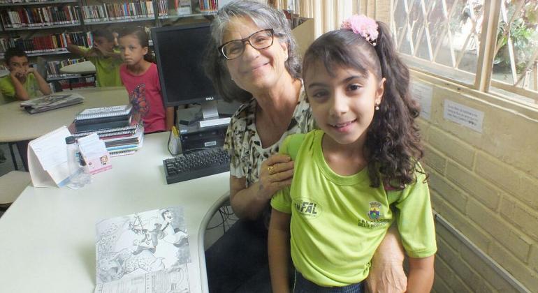 A Estudante Luísa Camargos, de 9 anos, se tornou escritora e está sentada no colo da bibliotecária, na biblioteca de sua escola. Ao fundo, dois colegas de Luísa estão sentados à mesa.