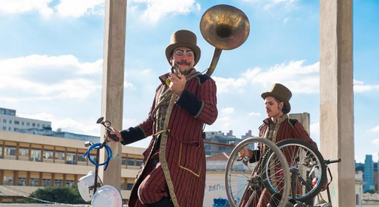 Dois atores em bicicletas, um deles com um trompete, ao ar livre.