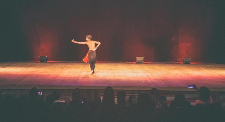 PBH divulga projetos habilitados e inabilitados na Lei de Incentivo à Cultura Garoto se apresenta no palco do Teatro Francisco Nunes usando uma calça preta e tecido vermelho amarrado na cintura.